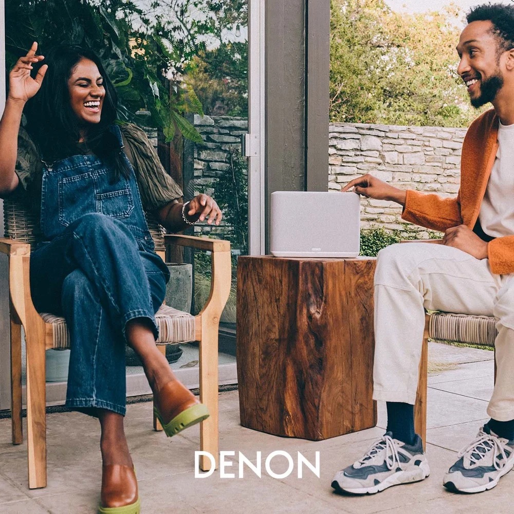 Two friends laughing on an outdoor patio, a Denon Home speaker sitting on a wood block between them; golden-hour light, garden and stone wall in the background.